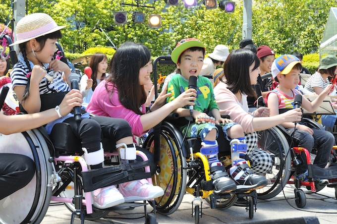 【写真】車椅子に乗る子どもたちにマイクを向けるスタッフ。子どもたちは楽しそうな表情をしていて、手には楽器を持っている。