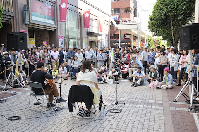 【写真】道路に椅子を置きギターを演奏し歌を歌っている人。周りを大勢の観客が囲み演奏を真剣に見つめている。