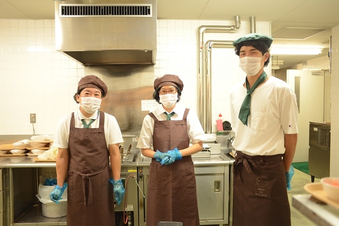 【写真】キッチンで働くスタッフさんたち。マスクとエプロンをしていながらも、笑みを浮かべていることがわかる。