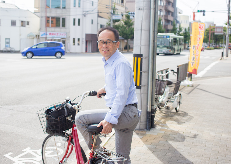【写真】街頭で笑顔で自転車に乗っているきもとつとむさん