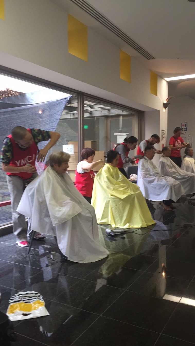 【写真】おちあいさんがヘアカットボランティアをしている様子。全部で５名の男女が一列に並び髪をカットしてもらっている。