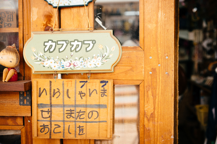 【写真】カプカプ の入り口にある木製の看板。手書きで「いらっしゃいませ、またのおこしを」と書いてある。