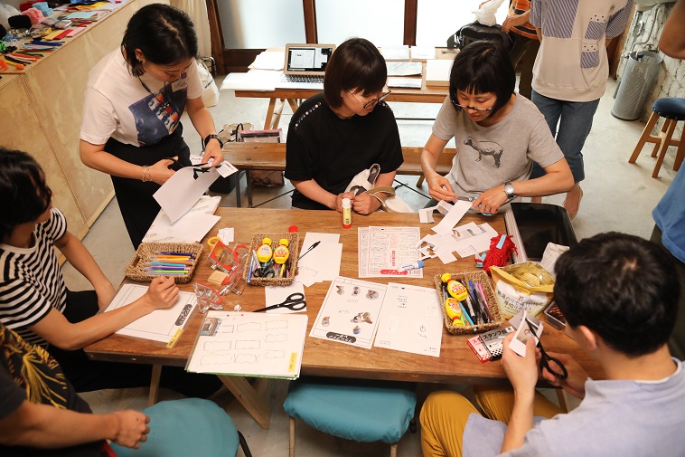 【写真】6人ほどの人が机を囲んでハサミやペンを使って作業をしている