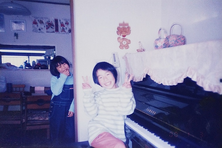 【写真】小学生の頃のあさのさん。ピアノの前に座り、両手でピースサインをしている。