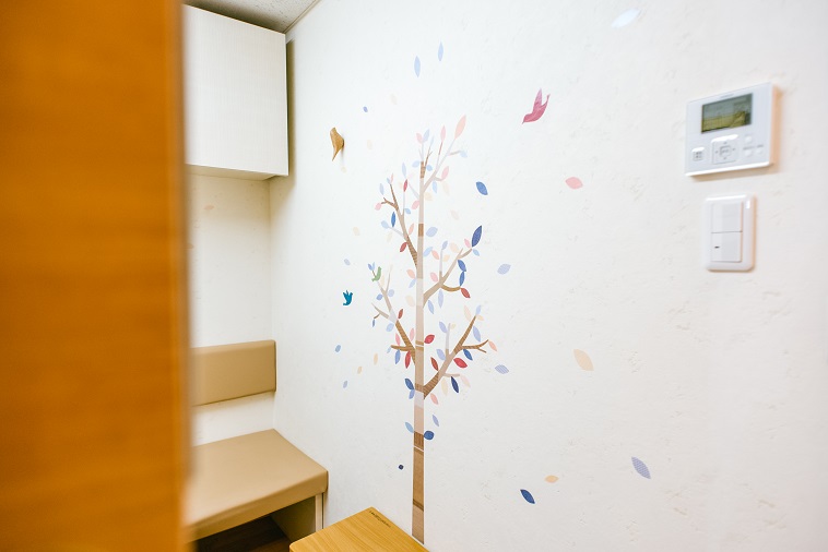 【写真】ロコクリニック中目黒の診察室。壁には大きな木に集まる鳥たちの絵がやわらかな色彩で描かれている。