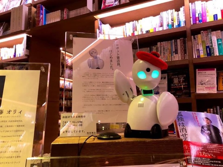 【写真】ベレー帽を被った卓上サイズのOriHimeが右腕を上げている。後ろには「ロボット書店員、はじめました。」と書かれた紙が