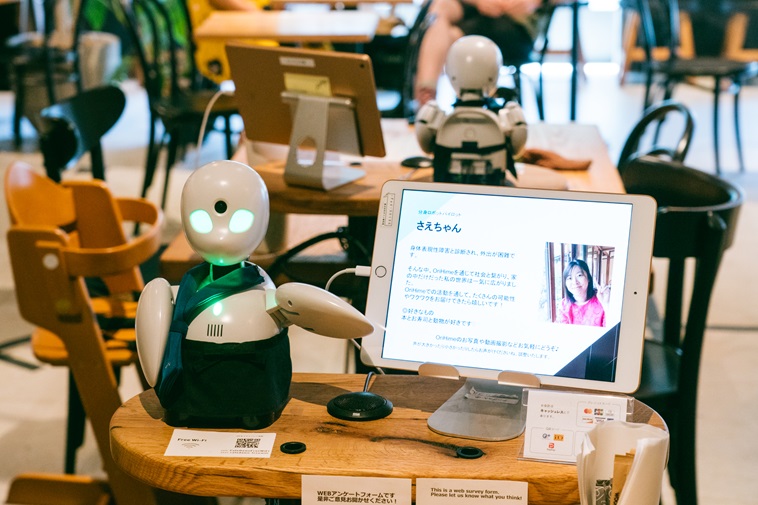 【写真】テーブルの上に20センチほどの白いロボットが座っている。目は光、左腕をあげる仕草をとっている。左腕の先には、さえさんの写真と自己紹介が書かれたiPad画面が。