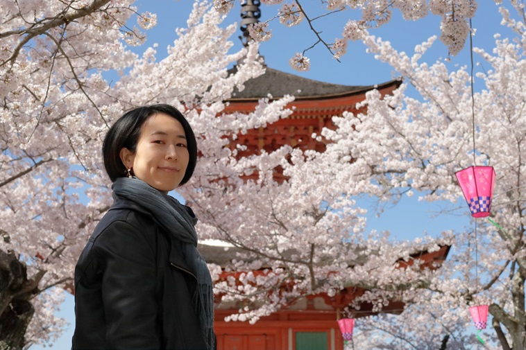 【写真】お寺と桜を背景にカメラを見つめるもりみさ