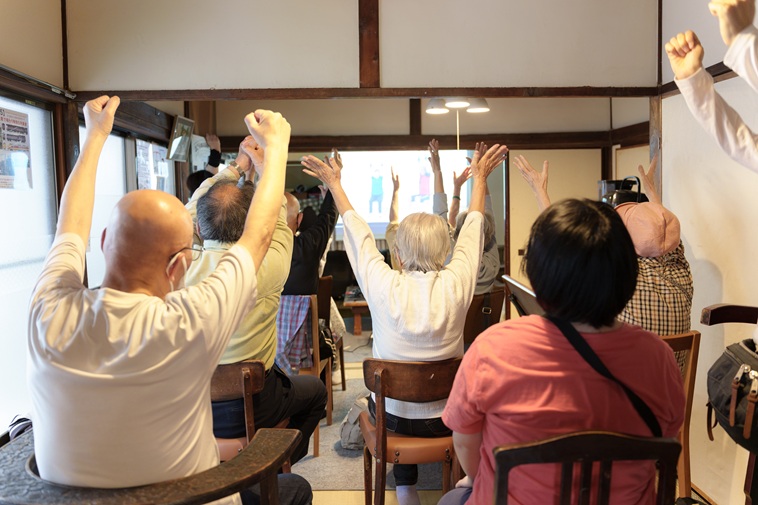 【写真】岡さんのいえの室内で椅子に座り、体操をする高齢者たち