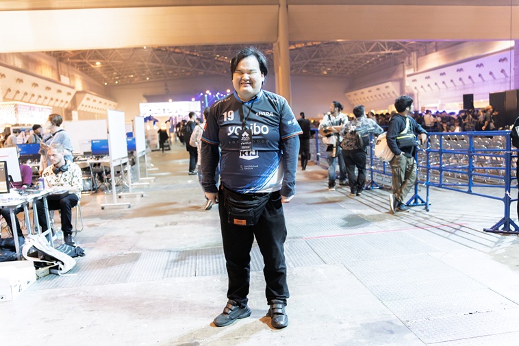 【写真】EVO Japanの会場をバックに、ePARAユニフォームを着てカメラに笑顔を向けるきたむらさん