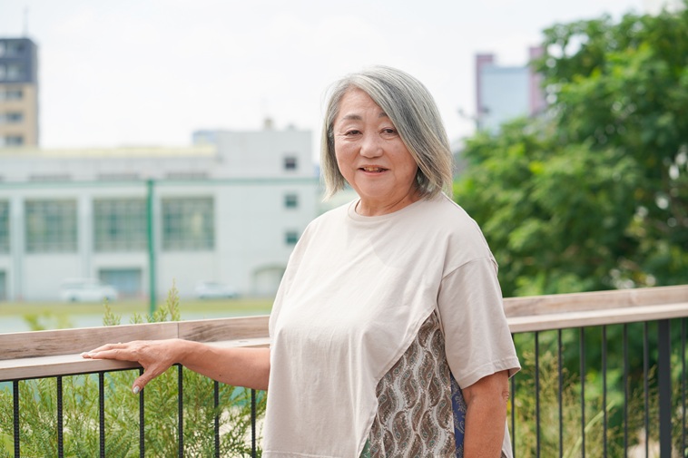 【写真】ベランダの手すりに手をかけカメラを見つめるよこやまとしこさん