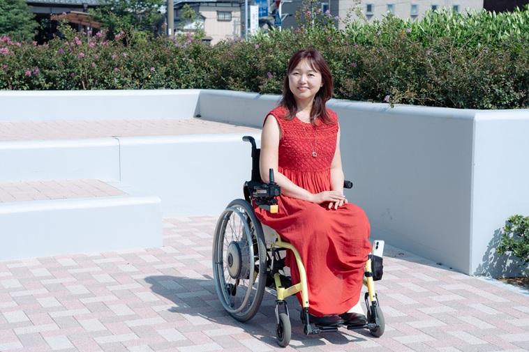 【写真】屋外で車いすに座り、微笑むおざわさん