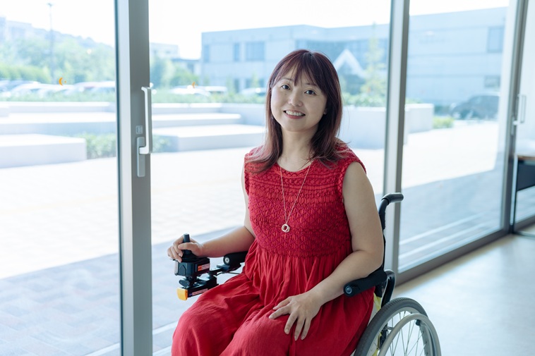 【写真】窓際で電動車いすに乗り微笑むおざわあやこさん