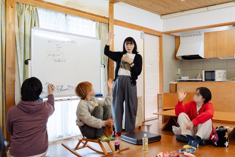 【写真】ホワイトボードの前に立つ江連麻紀さんと、円になって座っている参加者が手を挙げている