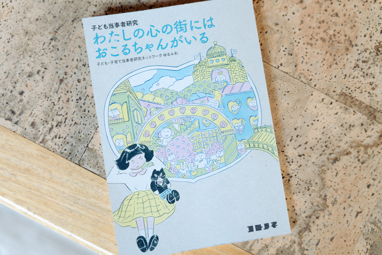 【写真】書籍『わたしの心の街にはおこるちゃんがいる』の表紙。女の子と猫のぬいぐるみ、笑顔の人がたくさんいる心の街のイメージのようなものが描かれている