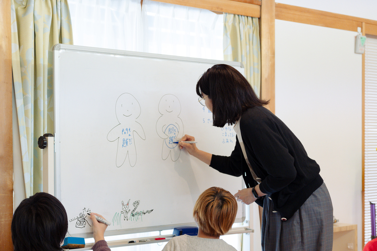 【写真】当事者研究中、子供と一緒にホワイトボードに絵を描いているえづれさん
