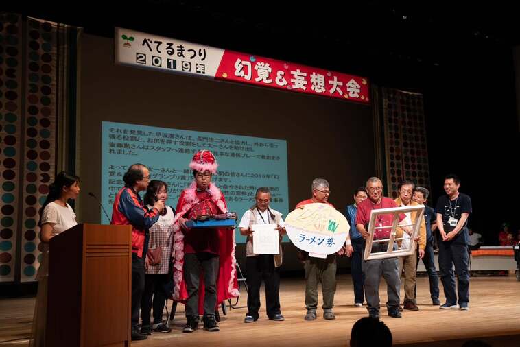 【写真】「べてるまつり2019年　幻覚＆妄想大会」と書かれた横断幕がかかったステージに、べてるの家の人たちが立って話をしている。器にラーメン券と書かれたラーメンの絵を持っている人、窓枠のようなものを持った人、赤いマントをつけた人など。