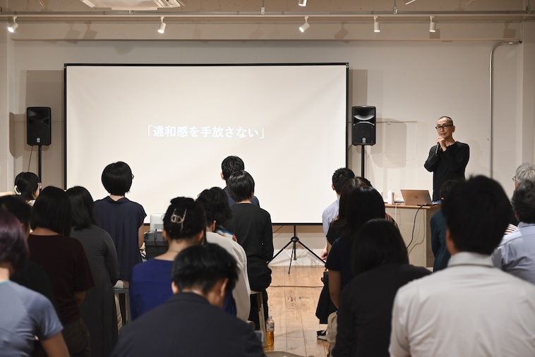 【写真】「違和感を手放さない」と書かれたスライドの横に立ち、マイクを握って話すにしむらさん