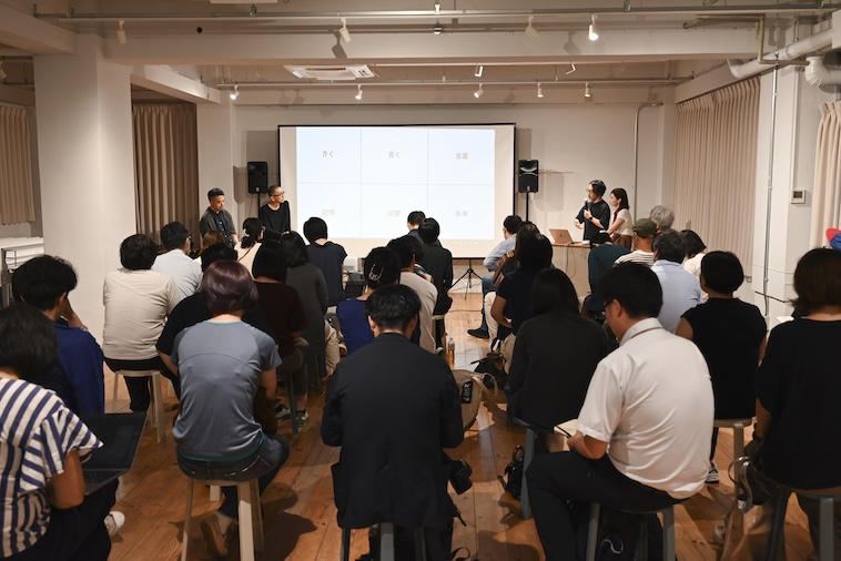【写真】会場を埋め尽くすたくさんの聴衆が前方のスライドを見ている